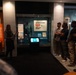 A museum guide is presenting an Olympic Games exhibition to a group of visitors. The display case features artifacts related to the Olympic Games, including a prominent sign reading 'Olympic Games Not War Games.' The exhibit appears to focus on the history of the Olympic Games, with visible references to Melbourne 1956 and Paris 1924.