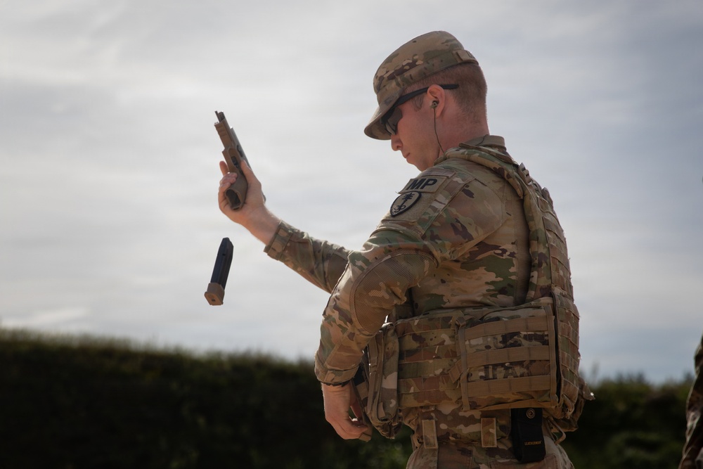 A military police soldier in OCP (Operational Camouflage Pattern) uniform is performing a magazine reload or weapons check with a pistol. The soldier has ejected or dropped a magazine while holding the pistol raised in their right hand. The image appears to be taken during a training exercise or qualification event.