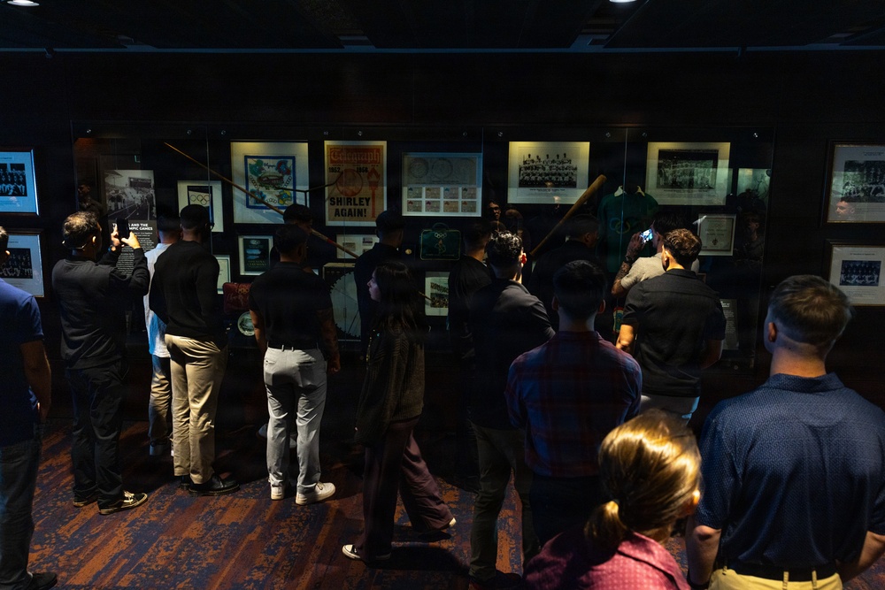 A group of visitors is viewing a sports memorabilia exhibition in what appears to be a museum or hall of fame. The display cases contain framed photographs, newspaper clippings, stamps, and sports artifacts including a prominent baseball bat and what appears to be athletic uniforms. One visible newspaper headline reads 'SHIRLEY AGAIN!' suggesting the exhibit may be related to a notable athlete named Shirley.
