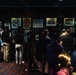 A group of visitors is viewing a sports memorabilia exhibition in what appears to be a museum or hall of fame. The display cases contain framed photographs, newspaper clippings, stamps, and sports artifacts including a prominent baseball bat and what appears to be athletic uniforms. One visible newspaper headline reads 'SHIRLEY AGAIN!' suggesting the exhibit may be related to a notable athlete named Shirley.