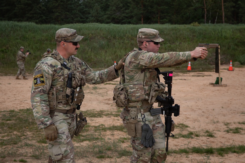 Two U.S. Army soldiers in MultiCam (OCP) uniforms are on a firing range, with one soldier appearing to coach or supervise the other who is aiming a pistol with an extended arm. Both soldiers are wearing full tactical gear including plate carriers, helmets/patrol caps, and carrying rifles slung on their bodies. Additional soldiers and orange traffic cones are visible in the background, indicating an organized range training event.