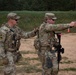 Two U.S. Army soldiers in MultiCam (OCP) uniforms are on a firing range, with one soldier appearing to coach or supervise the other who is aiming a pistol with an extended arm. Both soldiers are wearing full tactical gear including plate carriers, helmets/patrol caps, and carrying rifles slung on their bodies. Additional soldiers and orange traffic cones are visible in the background, indicating an organized range training event.