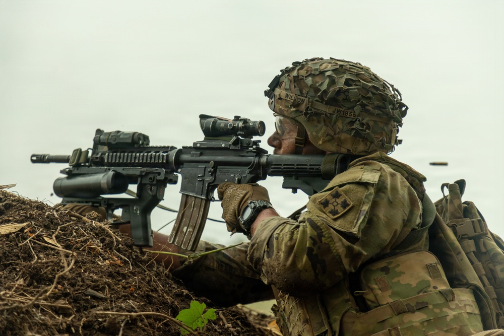 A U.S. Army soldier in full combat gear is positioned in a fighting trench or foxhole, aiming an M4 carbine rifle equipped with an optic scope and underbarrel attachment. The soldier wears OCP (Operational Camouflage Pattern) uniform with a unit patch visible on the shoulder and a camouflage-covered helmet. A spent brass casing is visible in the air to the right, indicating the weapon has recently been fired.