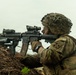 A U.S. Army soldier in full combat gear is positioned in a fighting trench or foxhole, aiming an M4 carbine rifle equipped with an optic scope and underbarrel attachment. The soldier wears OCP (Operational Camouflage Pattern) uniform with a unit patch visible on the shoulder and a camouflage-covered helmet. A spent brass casing is visible in the air to the right, indicating the weapon has recently been fired.