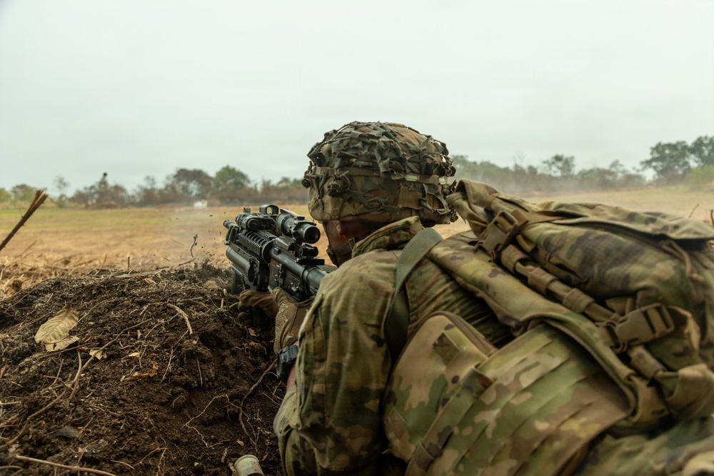 A soldier in full combat gear is positioned in a fighting hole or trench, aiming a rifle equipped with optical sights across an open field. The soldier is wearing multicam/OCP camouflage uniform with a helmet covered in camouflage netting and carrying a large rucksack. The setting appears to be a military training exercise or field operation in a semi-arid, open terrain environment.