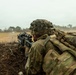 A soldier in full combat gear is positioned in a fighting hole or trench, aiming a rifle equipped with optical sights across an open field. The soldier is wearing multicam/OCP camouflage uniform with a helmet covered in camouflage netting and carrying a large rucksack. The setting appears to be a military training exercise or field operation in a semi-arid, open terrain environment.