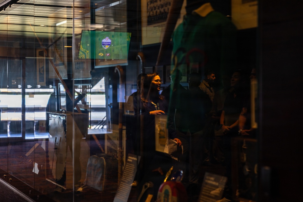 The image shows people inside what appears to be a sports bar or venue, viewed through a glass display case or window. A woman wearing glasses is prominently visible in the center, with several other individuals standing behind her. A television screen displaying what appears to be a sports broadcast is visible in the background.