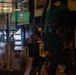 The image shows people inside what appears to be a sports bar or venue, viewed through a glass display case or window. A woman wearing glasses is prominently visible in the center, with several other individuals standing behind her. A television screen displaying what appears to be a sports broadcast is visible in the background.