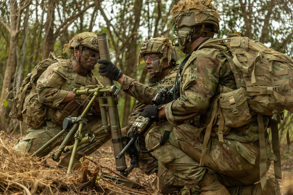 Three soldiers in full combat gear are gathered around what appears to be a mortar system in a wooded outdoor environment. One soldier appears to be instructing or directing the others in the setup or operation of the weapon system. All personnel are wearing camouflage uniforms, helmets with foliage, face paint, and tactical backpacks.