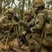 Three soldiers in full combat gear are gathered around what appears to be a mortar system in a wooded outdoor environment. One soldier appears to be instructing or directing the others in the setup or operation of the weapon system. All personnel are wearing camouflage uniforms, helmets with foliage, face paint, and tactical backpacks.