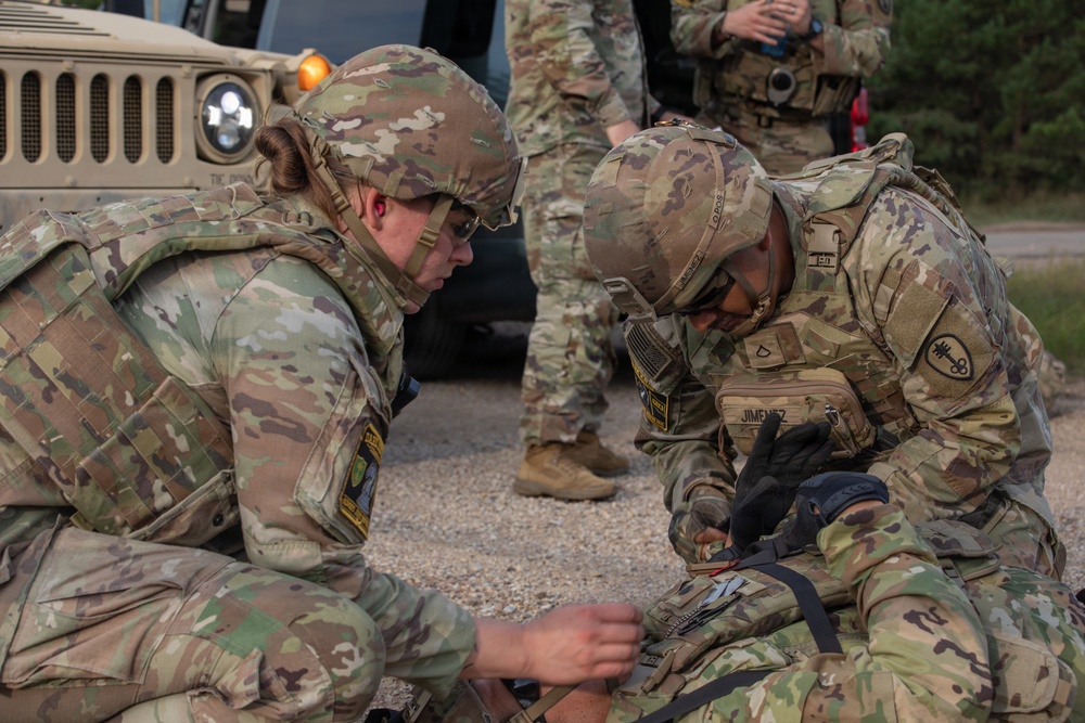 Two U.S. Army soldiers in OCP (Operational Camouflage Pattern) uniforms are kneeling on gravel, performing what appears to be a tactical combat casualty care (TCCC) or first aid training exercise. One soldier, identified by a name tape reading 'JIMENEZ,' is focused on applying what appears to be a tourniquet or medical treatment to a simulated casualty. Additional personnel and a military HMMWV (Humvee) vehicle are visible in the background.