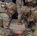 Two U.S. Army soldiers in OCP (Operational Camouflage Pattern) uniforms are kneeling on gravel, performing what appears to be a tactical combat casualty care (TCCC) or first aid training exercise. One soldier, identified by a name tape reading 'JIMENEZ,' is focused on applying what appears to be a tourniquet or medical treatment to a simulated casualty. Additional personnel and a military HMMWV (Humvee) vehicle are visible in the background.