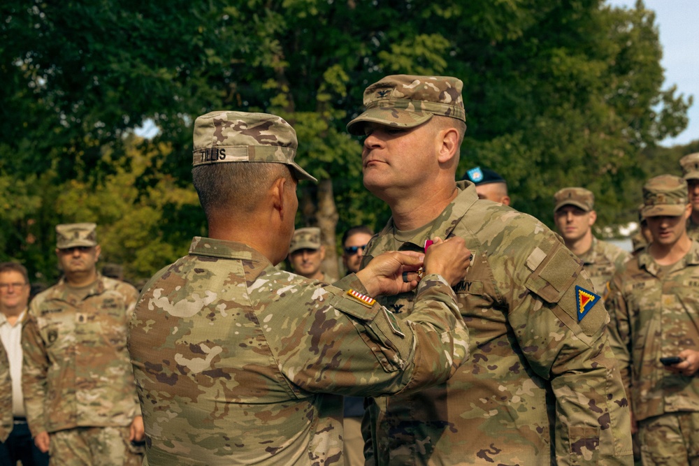 A military medal pinning ceremony is taking place outdoors, with one soldier pinning a medal onto the chest of another soldier of higher rank. The recipient wears a colonel's eagle rank insignia on his cap and displays a unit patch on his left shoulder. Multiple uniformed soldiers stand in formation as witnesses in the background.