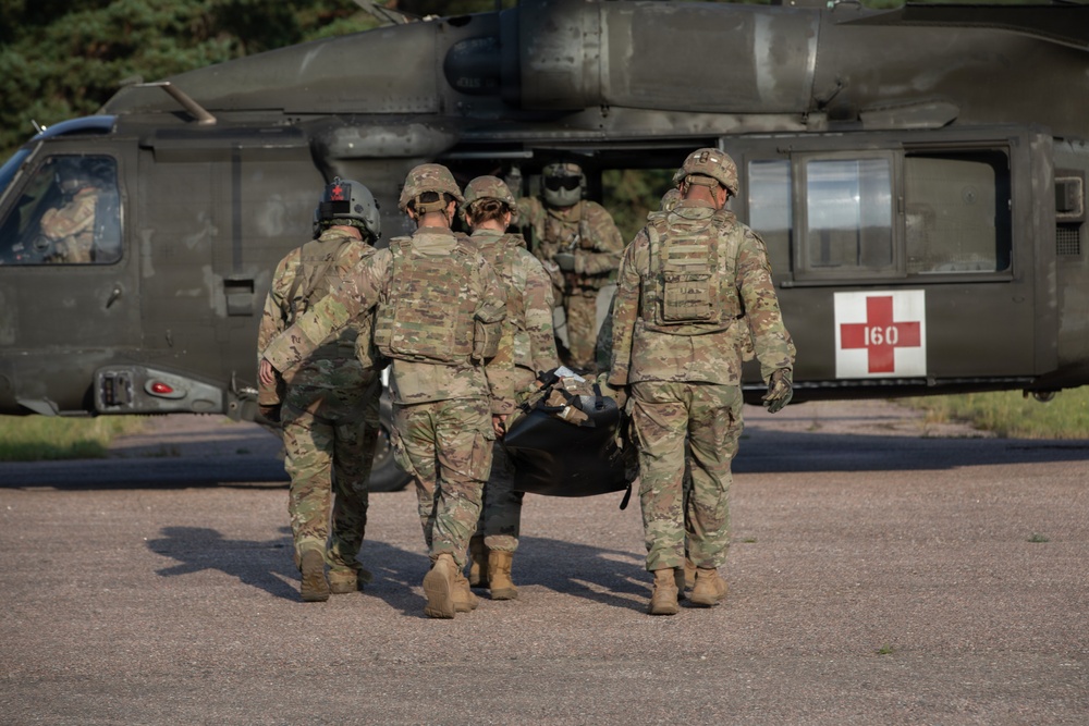 Military personnel in camouflage uniforms are carrying a patient on a litter/stretcher toward a military medevac helicopter marked with a red cross and the number '160'. The helicopter appears to be a UH-60 Black Hawk configured for medical evacuation (MEDEVAC). This appears to be either a training exercise or an actual casualty evacuation operation.
