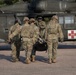 Military personnel in camouflage uniforms are carrying a patient on a litter/stretcher toward a military medevac helicopter marked with a red cross and the number '160'. The helicopter appears to be a UH-60 Black Hawk configured for medical evacuation (MEDEVAC). This appears to be either a training exercise or an actual casualty evacuation operation.
