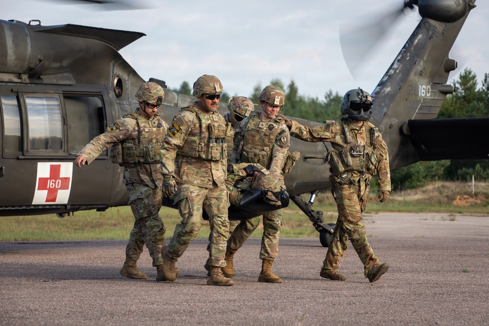 Military personnel in camouflage uniforms are carrying a litter/stretcher away from a UH-60 Black Hawk medical evacuation helicopter marked with a red cross and the number 160. The scene appears to depict a MEDEVAC training exercise or casualty evacuation operation on a tarmac or airfield. Four soldiers are visible, including what appears to be a flight crew member wearing a flight helmet.