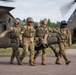 Military personnel in camouflage uniforms are carrying a litter/stretcher away from a UH-60 Black Hawk medical evacuation helicopter marked with a red cross and the number 160. The scene appears to depict a MEDEVAC training exercise or casualty evacuation operation on a tarmac or airfield. Four soldiers are visible, including what appears to be a flight crew member wearing a flight helmet.