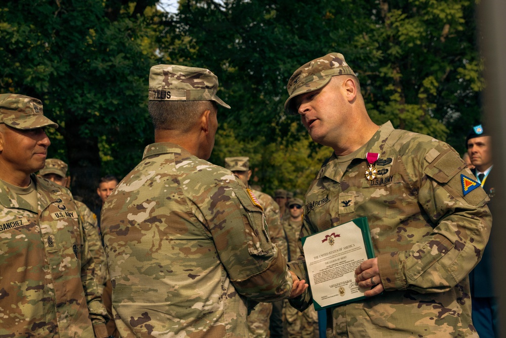 A U.S. Army military ceremony is taking place outdoors, where a Colonel (identified by eagle insignia) is receiving or presenting a Legion of Merit award. Two soldiers are shaking hands while the Colonel holds the award certificate, with the Legion of Merit medal visibly pinned to his uniform. Additional soldiers and personnel are visible in the background.
