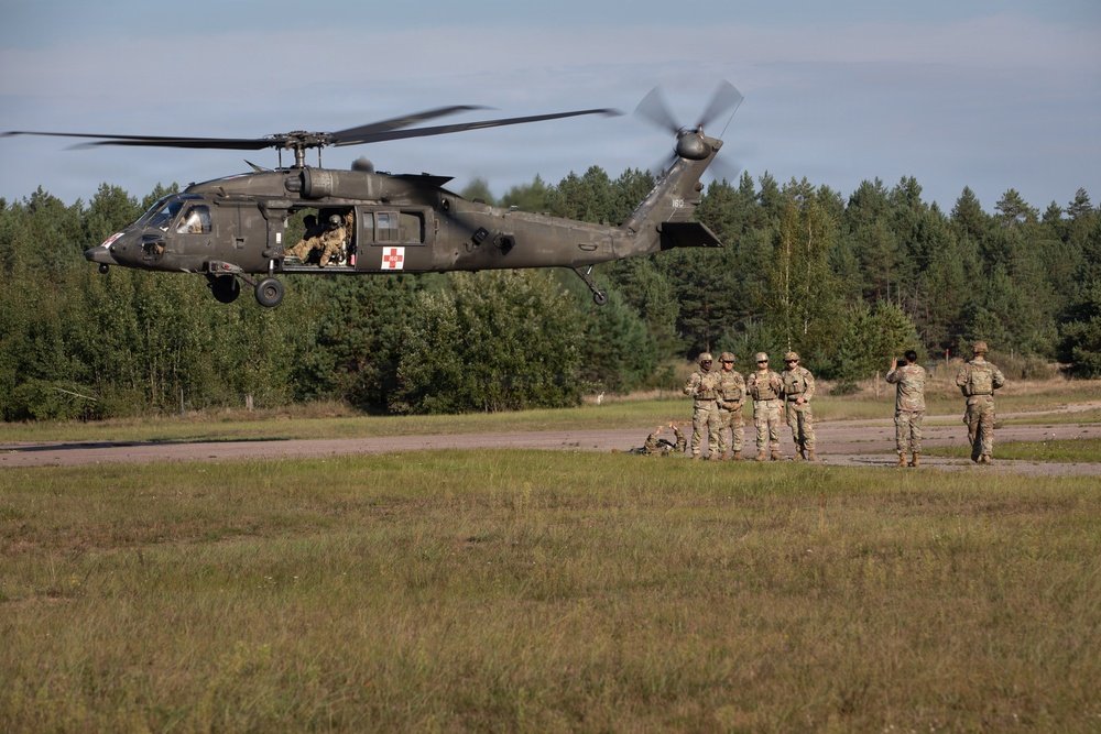 A U.S. Army UH-60 Black Hawk helicopter marked with a red cross medical insignia hovers low over a grassy landing zone. Several soldiers in camouflage uniforms stand on the ground, with at least one appearing to signal the aircraft. The scene appears to depict a medical evacuation (MEDEVAC) training exercise in a forested area.