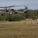A U.S. Army UH-60 Black Hawk helicopter marked with a red cross medical insignia hovers low over a grassy landing zone. Several soldiers in camouflage uniforms stand on the ground, with at least one appearing to signal the aircraft. The scene appears to depict a medical evacuation (MEDEVAC) training exercise in a forested area.