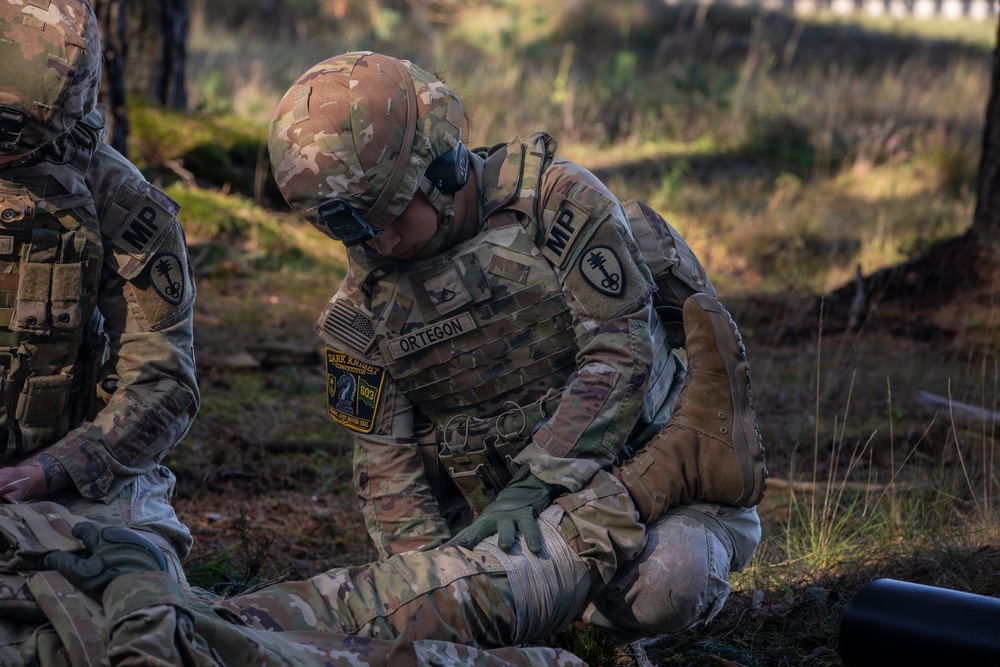 A U.S. Army Military Police soldier named Ortegon, assigned to the 603rd unit ('Dark Knights'), is performing what appears to be a tactical combat casualty care (TCCC) procedure on a simulated or actual casualty on the ground. A second MP soldier is visible on the left side of the frame. The setting appears to be an outdoor field training environment.