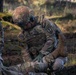 A U.S. Army Military Police soldier named Ortegon, assigned to the 603rd unit ('Dark Knights'), is performing what appears to be a tactical combat casualty care (TCCC) procedure on a simulated or actual casualty on the ground. A second MP soldier is visible on the left side of the frame. The setting appears to be an outdoor field training environment.