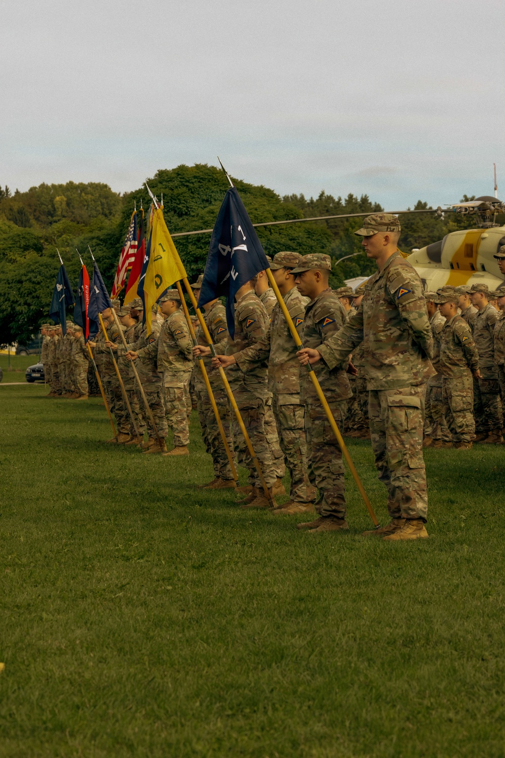 A formal U.S. Army military ceremony is taking place on a grass field, with soldiers in OCP (Operational Camouflage Pattern) uniforms holding unit guidons and flags in a ceremonial formation. Multiple guidon bearers are visible holding flags including blue, yellow, and red pennants, along with what appears to be a U.S. national flag. A military helicopter (appearing yellow and grey) is visible in the background, suggesting this may be an aviation unit ceremony.