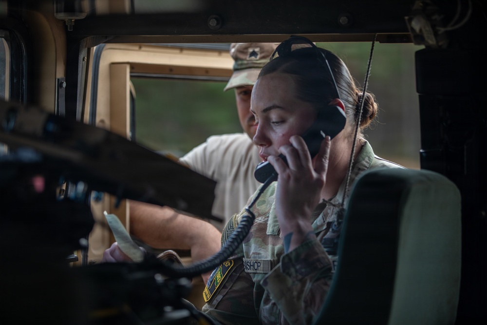 A female soldier with the name tape 'BISHOP' is seated inside a military vehicle, likely a HMMWV (Humvee), using a radio handset to communicate. She is wearing camouflage uniform with visible unit patches and a headset. A second individual in a white t-shirt and camouflage cap is visible in the background, appearing to observe or assist.