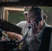 A female soldier with the name tape 'BISHOP' is seated inside a military vehicle, likely a HMMWV (Humvee), using a radio handset to communicate. She is wearing camouflage uniform with visible unit patches and a headset. A second individual in a white t-shirt and camouflage cap is visible in the background, appearing to observe or assist.
