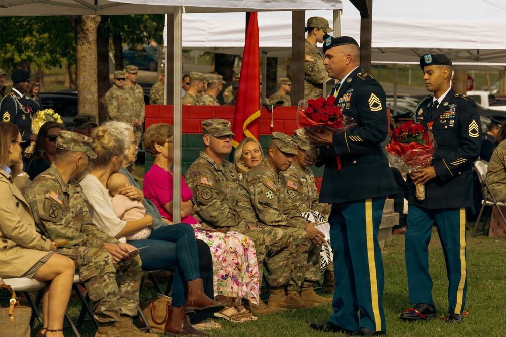 A U.S. Army military ceremony is taking place outdoors, likely a retirement or awards ceremony. Two soldiers in dress blue uniforms are holding bouquets of red roses, standing before an audience of seated soldiers in combat uniforms and civilian family members. A red unit guidon flag is visible in the background under a white canopy tent.