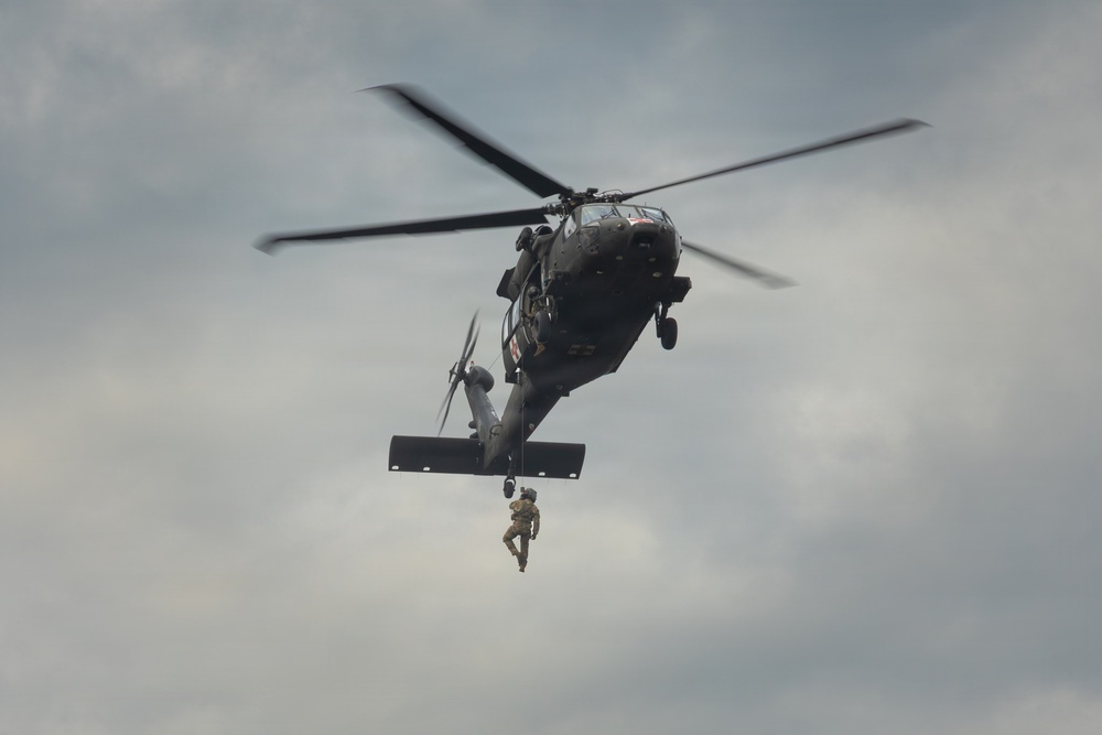 A military helicopter, consistent with a UH-60 Black Hawk, is hovering in an overcast sky while a soldier rappels or is being hoisted beneath it. The aircraft is dark/olive drab in color and appears to be conducting a training or operational insertion/extraction exercise. The rotor blades show motion blur, indicating active flight.