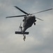 A military helicopter, consistent with a UH-60 Black Hawk, is hovering in an overcast sky while a soldier rappels or is being hoisted beneath it. The aircraft is dark/olive drab in color and appears to be conducting a training or operational insertion/extraction exercise. The rotor blades show motion blur, indicating active flight.