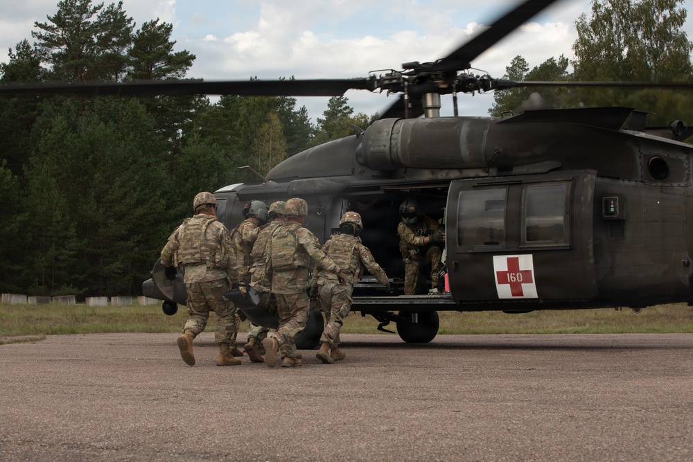Military personnel in camouflage uniforms are loading a casualty on a litter into a medical evacuation (MEDEVAC) helicopter. The helicopter, marked with a red cross and the number '160', is a UH-60 Black Hawk configured for medical evacuation. This appears to be a MEDEVAC training exercise or an actual casualty evacuation operation on a paved landing zone surrounded by forest.