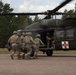 Military personnel in camouflage uniforms are loading a casualty on a litter into a medical evacuation (MEDEVAC) helicopter. The helicopter, marked with a red cross and the number '160', is a UH-60 Black Hawk configured for medical evacuation. This appears to be a MEDEVAC training exercise or an actual casualty evacuation operation on a paved landing zone surrounded by forest.