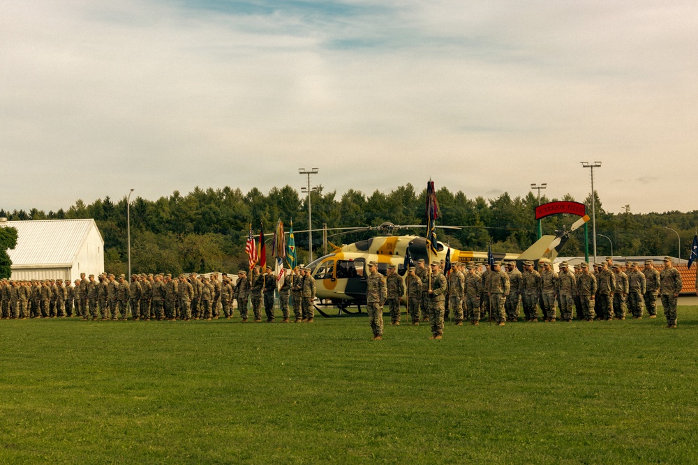 A military formation ceremony is taking place on a large grass field, with dozens of soldiers in camouflage uniforms standing in organized rows. A yellow and tan camouflage-painted helicopter is positioned centrally behind the formation, serving as a backdrop. Multiple military flags and guidons are visible, suggesting a formal unit ceremony such as a change of command or activation event.