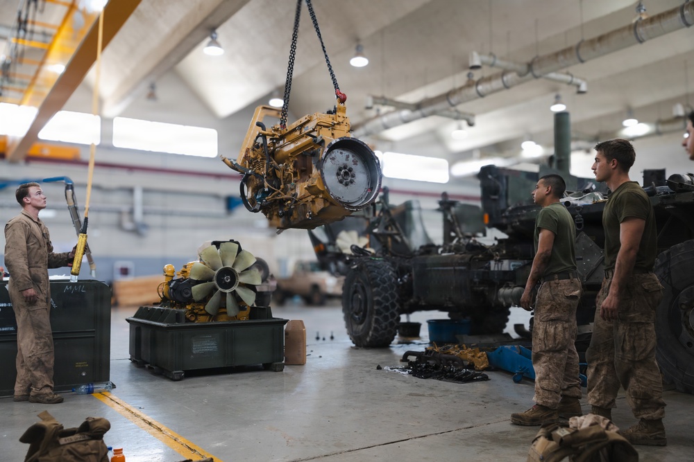 Military personnel are conducting heavy equipment maintenance inside a large vehicle repair bay or motor pool facility. A large yellow diesel engine is suspended by a chain hoist/overhead crane, being lifted or positioned during an engine swap or removal procedure. At least three service members in military uniforms are observing and supervising the lift operation.