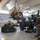 Military personnel are conducting heavy equipment maintenance inside a large vehicle repair bay or motor pool facility. A large yellow diesel engine is suspended by a chain hoist/overhead crane, being lifted or positioned during an engine swap or removal procedure. At least three service members in military uniforms are observing and supervising the lift operation.