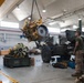 Military personnel are conducting heavy equipment maintenance inside a large vehicle repair bay or motor pool facility. A large yellow diesel engine is suspended by a chain hoist/overhead crane, being lifted or positioned during an engine swap or removal procedure. At least three service members in military uniforms are observing and supervising the lift operation.