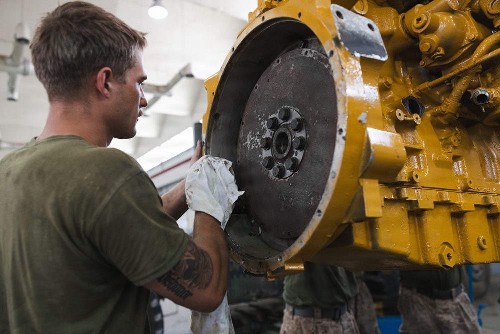 A young male mechanic wearing a green t-shirt and work gloves is inspecting or servicing a large yellow industrial engine or heavy equipment component. He is closely examining a large flywheel or clutch plate attached to the yellow-painted machinery. The work is taking place in what appears to be an indoor maintenance or repair facility.