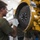 A young male mechanic wearing a green t-shirt and work gloves is inspecting or servicing a large yellow industrial engine or heavy equipment component. He is closely examining a large flywheel or clutch plate attached to the yellow-painted machinery. The work is taking place in what appears to be an indoor maintenance or repair facility.