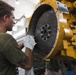 A young male mechanic wearing a green t-shirt and work gloves is inspecting or servicing a large yellow industrial engine or heavy equipment component. He is closely examining a large flywheel or clutch plate attached to the yellow-painted machinery. The work is taking place in what appears to be an indoor maintenance or repair facility.