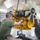 Military personnel in a maintenance bay are working on a large yellow diesel engine suspended by chains from an overhead hoist. At least two service members in military-style clothing are closely inspecting and handling the engine, which appears to be a Caterpillar-type industrial or heavy vehicle engine. The setting is an indoor workshop or motor pool facility with high ceilings and overhead lighting.