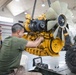 Military personnel in a maintenance bay are working on a large yellow diesel engine suspended by chains from an overhead hoist. At least two service members in military-style clothing are closely inspecting and handling the engine, which appears to be a Caterpillar-type industrial or heavy vehicle engine. The setting is an indoor workshop or motor pool facility with high ceilings and overhead lighting.