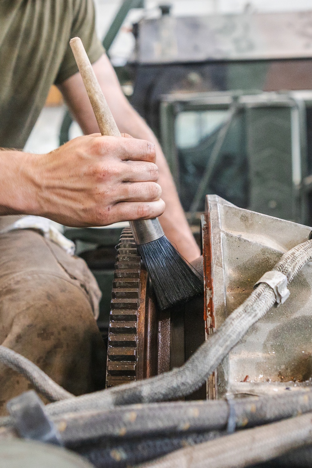 A mechanic or technician is using a large paintbrush-style brush with a wooden handle to clean or apply lubricant to a large industrial gear or sprocket. The machinery appears heavily worn and dirty, consistent with industrial or heavy equipment maintenance. The worker is wearing work-stained clothing, indicating active hands-on mechanical work.