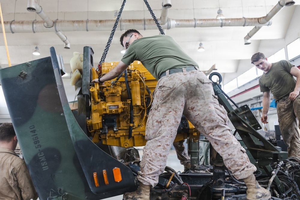U.S. Marine Corps personnel are performing engine maintenance or replacement in a military repair facility. A bright yellow engine block is being lowered by chain hoist into a piece of green military equipment. The personnel are wearing USMC desert camouflage (MARPAT) uniforms and olive drab t-shirts.