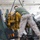 U.S. Marine Corps personnel are performing engine maintenance or replacement in a military repair facility. A bright yellow engine block is being lowered by chain hoist into a piece of green military equipment. The personnel are wearing USMC desert camouflage (MARPAT) uniforms and olive drab t-shirts.