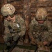 Two U.S. Army soldiers in Operational Camouflage Pattern (OCP) uniforms and helmets are working inside a log structure, focused on equipment or a task on what appears to be a litter or stretcher. The soldier on the right has 'MP' (Military Police) markings visible on their chest. A third person in civilian/non-combat attire is partially visible on the left edge of the frame.