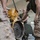 Military personnel in camouflage uniforms are working together in a maintenance bay to handle a large yellow engine or mechanical assembly suspended by a chain hoist. The team appears to be positioning or installing the heavy component, which shows a visible flywheel and bell housing. The setting is an indoor workshop or motor pool facility.