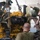 Two military personnel in camouflage uniforms are working on a large yellow industrial engine suspended by chains in what appears to be a maintenance bay or motor pool facility. One service member is leaning over the engine handling a fan blade component while the other works from below. The engine appears to be a Caterpillar-type diesel engine undergoing installation or removal.