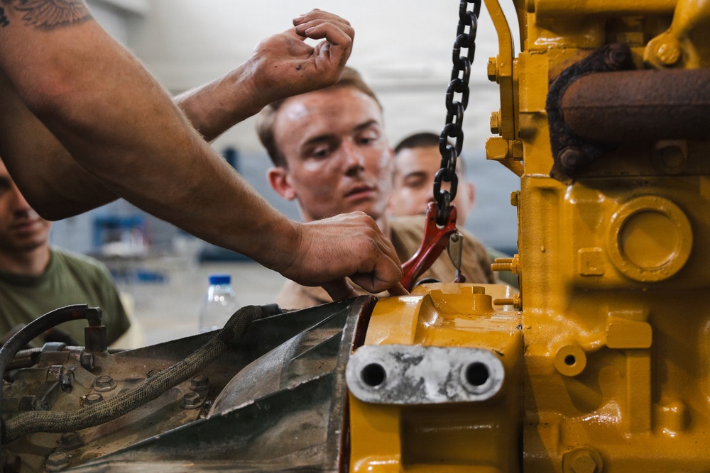 Several individuals, appearing to be military personnel or trainees in a workshop setting, are working on a large yellow industrial or military engine component. A chain hoist with a red hook is being used to assist with the mechanical work. The scene suggests a hands-on maintenance training exercise.
